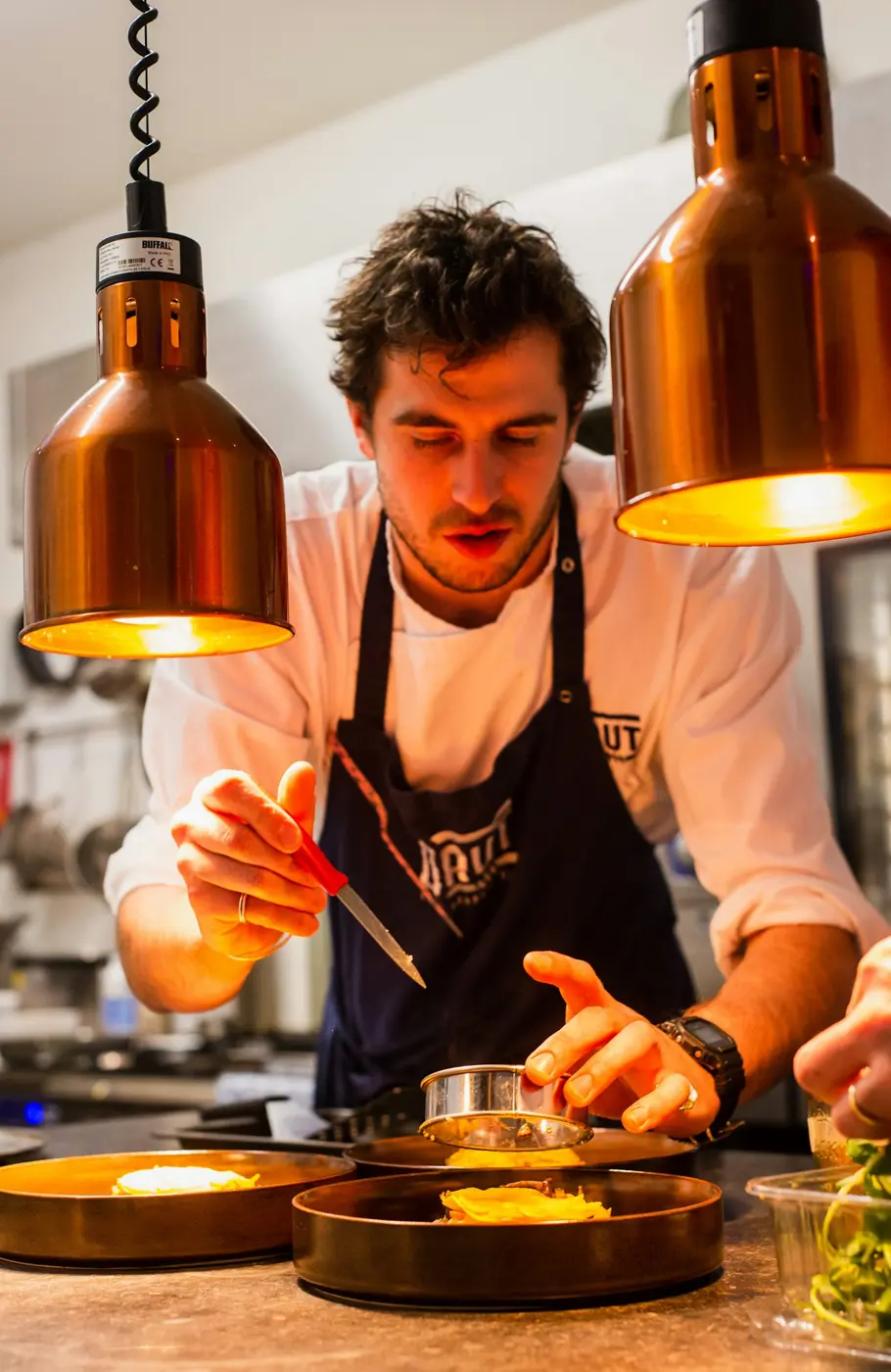 A chef plating a dish under heat lamps in a busy kitchen